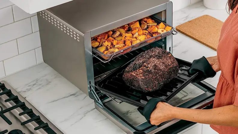 Person pulling pork roast out of Ninja oven, with roasted potatoes on the top shelf