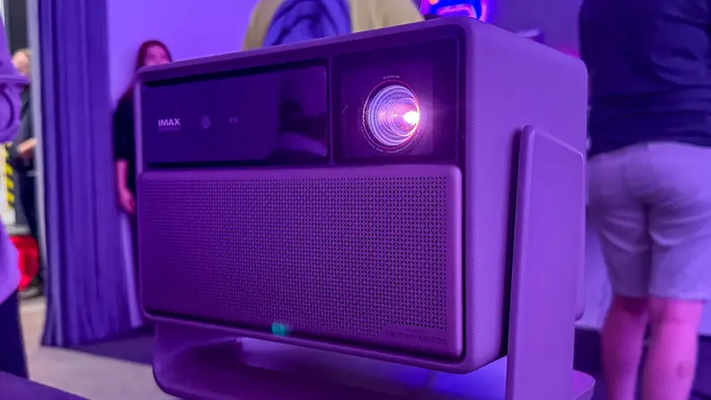 A close-up of an XGIMI Horizon 20 series projector is shown on display, its lens glowing with purple light in a dimly lit room. The projector features a perforated speaker grille while people stand blurred in the background.