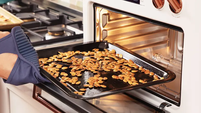 A person pulling out a metal sheet with baked pumpkin seeds on it, out of the oven.