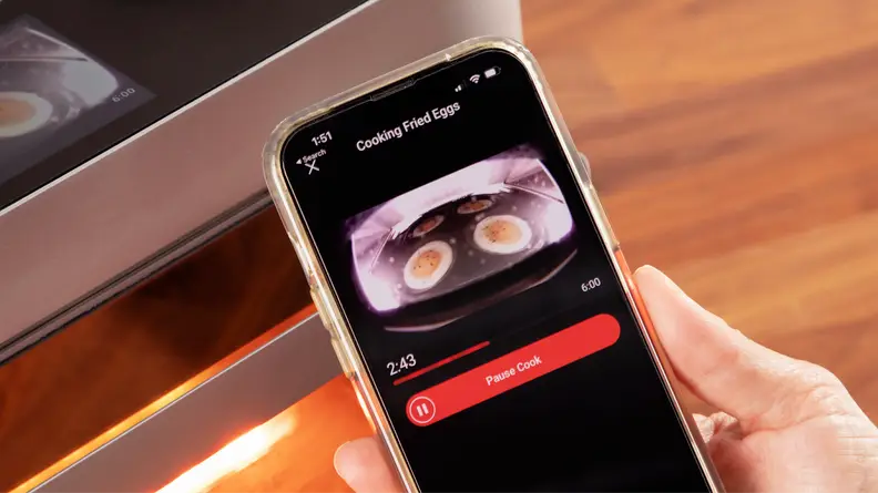 A person holding an iPhone in their hand with eggs cooking inside of the Brava Glass oven on screen.