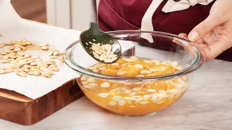 A person holding a clear bowl of pumpkin seeds and scooping pumpkin seeds out of the bowl with the black spoon.