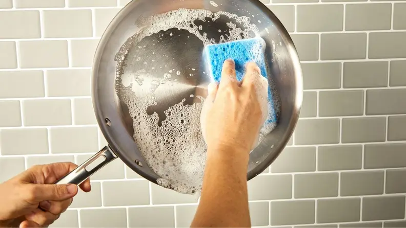 A person scrubs a stainless steel skillet with a sponge.