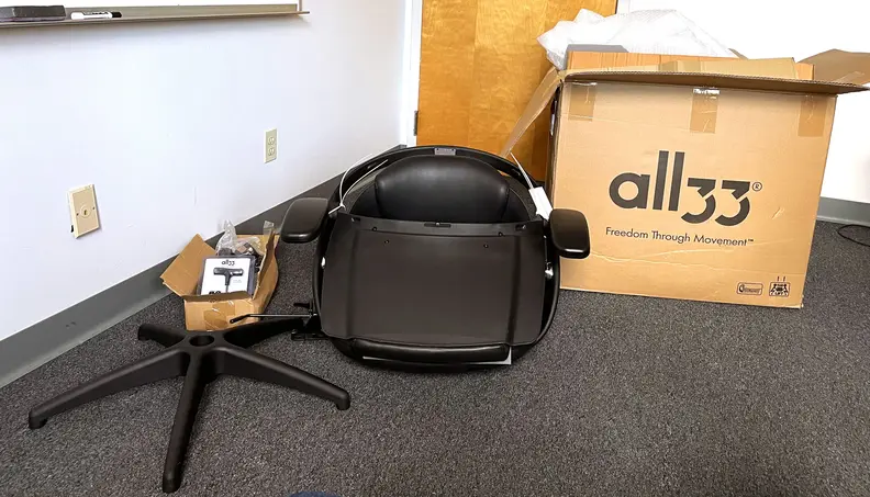 In the corner of a conference room you can see an opened All33-branded box. To its side are the main components that came inside: the chair itself, which is folded up, the bottom legs, a bracket for adjusting height, and a small box that holds the wheels, hydraulic lift, screws, and hex key.