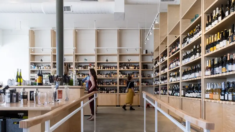 Four people shop for wine in a brightly lit natural wine store with light wood shelves filled with colorful bottles.