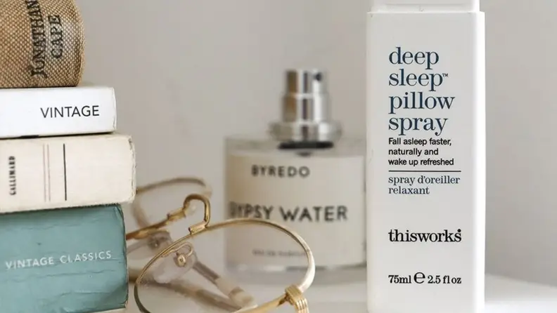 the Thisworks pillow spray sits on a side table next to glasses and books