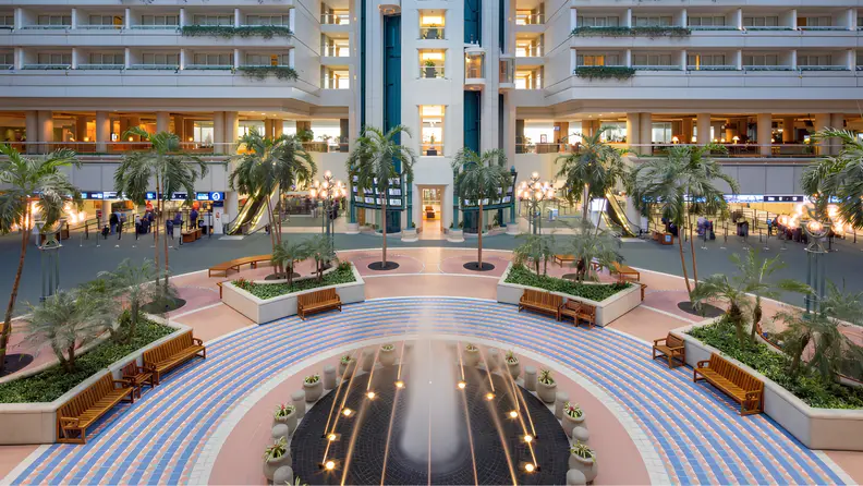 Spacious hotel atrium with indoor palm trees, a central fountain, and open seating areas surrounded by multiple floors of balconies and escalators.