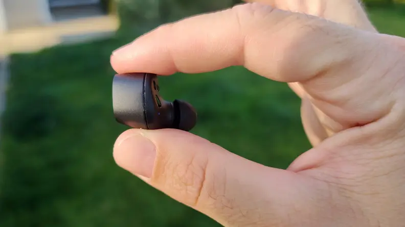 One Sennheiser CX Plus all-black earbud being held between two fingers in front of a lawn.