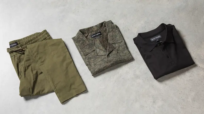 A pair of green pants, a camouflage camp shirt, and a black polo on a tabletop.