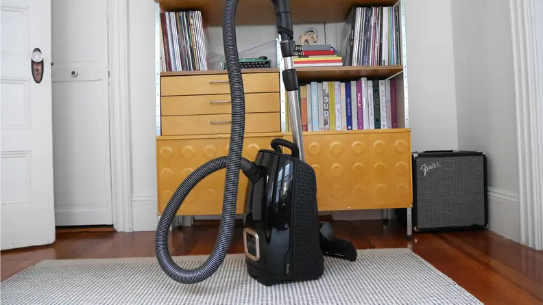 Miele Guard L1 Cat & Dog Vacuum standing up on a white and grey rug in front of a wooden cabinet with drawers and shelves with a white wall behind it.