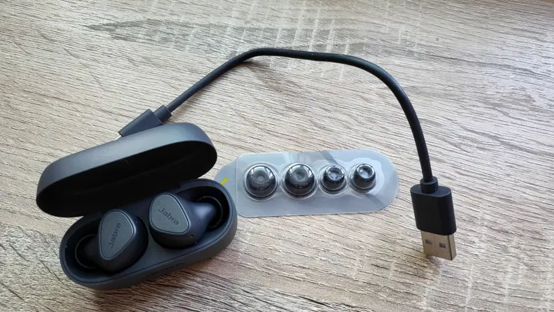 Jabra's all-black, matte earbuds sit in their open case on a wooden surface next to a small USB charing cable and a package of silicone eartips.