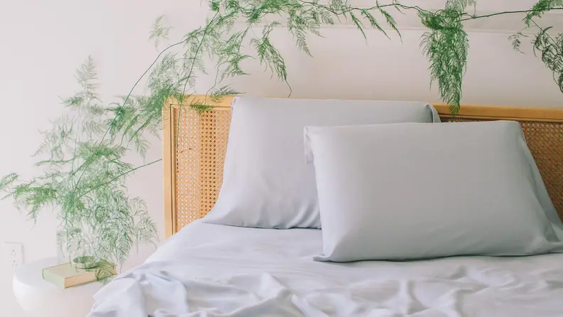 Wooden bed fitted with light grey sheets with plant on bedside table