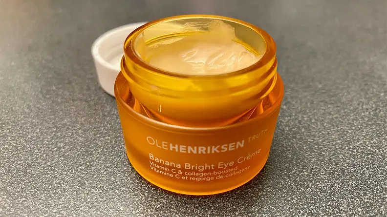 The OleHenriksen Banana Bright Eye Crème sitting on a bathroom vanity with its lid off to expose a orange-tinted cream.