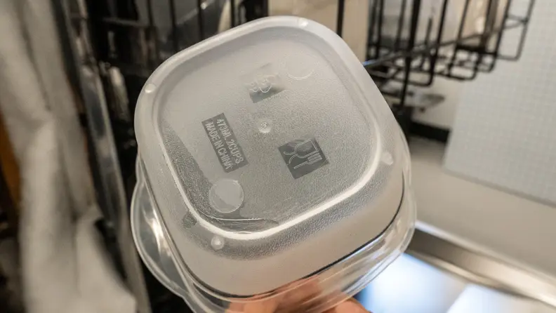 A hand holds a reusable plastic food container up to the camera, in front of the GE Café CDT875M5NS5 dishwasher. It looks bone dry.