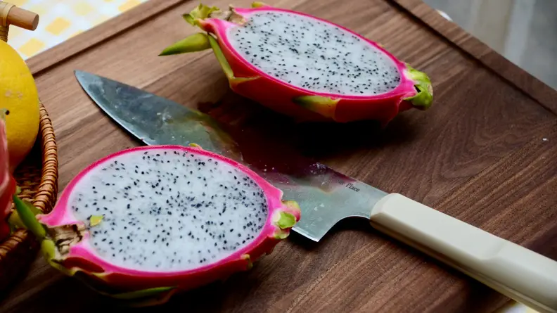 On an Our Place walnut wood cutting board, there are two halves of a dragon fruit. Between the halves, there's an Our Place chef's knife.