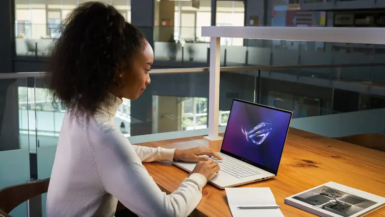 A girl sits in profile in front of an Asus ROG Zephyrus on a desk in a public setting.