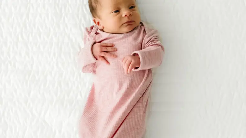 Newborn baby in a pink gown lying awake on a white textured blanket.