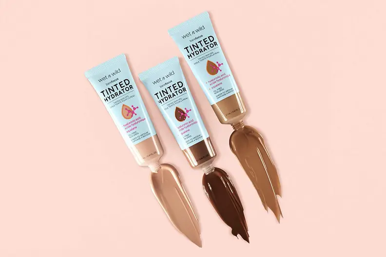 Three Wet n Wild Bare Focus Tinted Hydrators in light, medium, and dark skin tones lay on a light pink background with swatches of the formula coming out of the tubes.