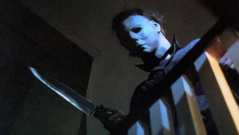 Michael Myers with a butcher knife leering over a staircase railing.