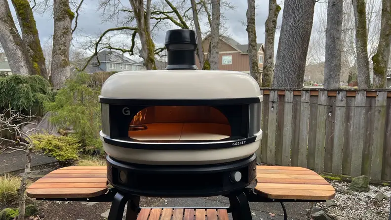 Gozney Dome Pizza Oven on stand with wood side tables, outdoors with open flame inside.