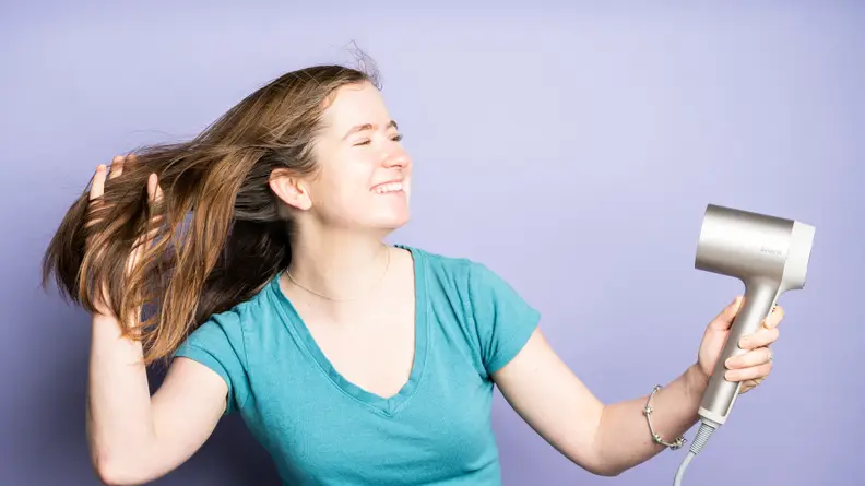 A person wearing a blue shirt standing against a purple background pointing a Shark hair dryer at their long brown hair.