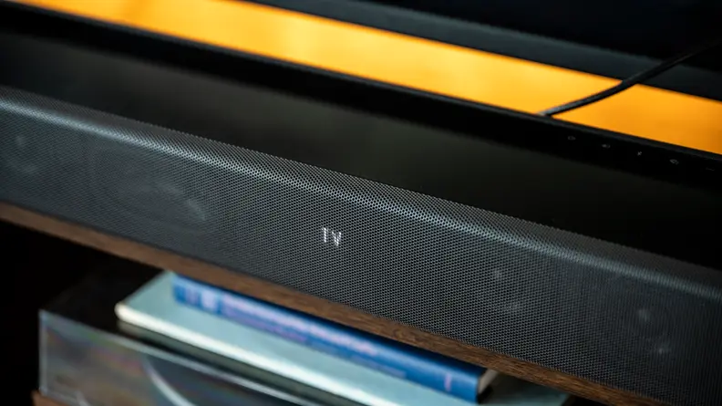 The all black Sony HT-A7000 sits atop a dark wood console against a brick wall below a large flat screen TV showing its LED display.
