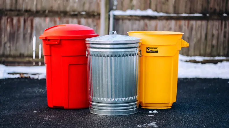 A red trash can, silver steel trash can, and yellow trash can sit on asphalt in front of a wooden fence.