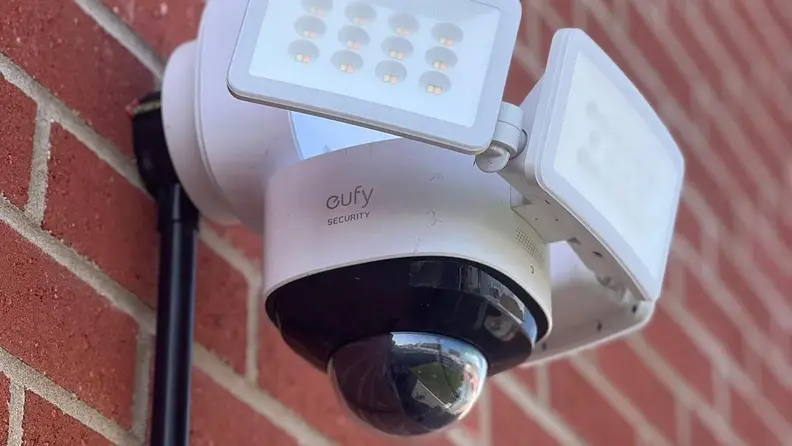 White surveillance system with built-in cameras and lights mounted on brick wall.