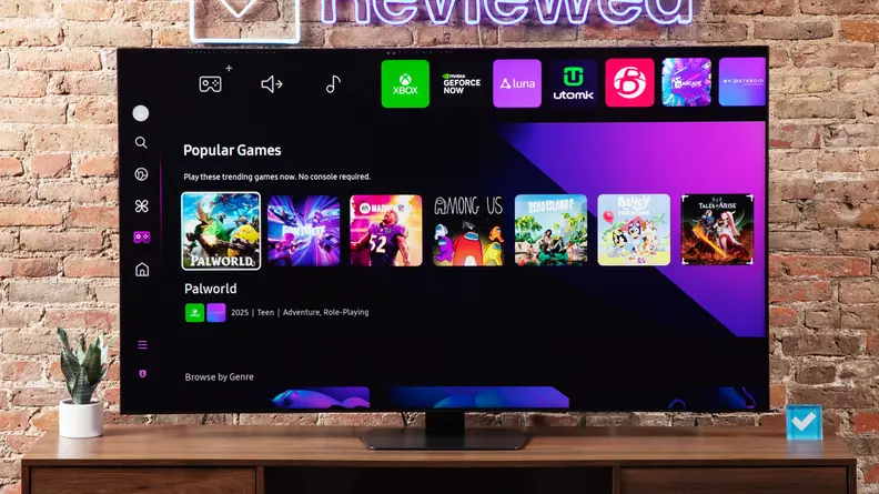Samsung QN90D Mini-LED TV with Gaming Hub on screen atop a wooden TV stand below Reviewed neon sign in front of brick wall indoors.