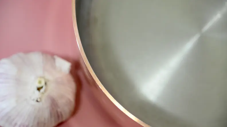 A close-up photo of a copper pan with a garlic bulb next to it on a pink background.