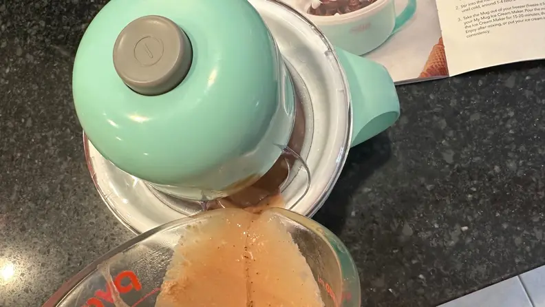 A measuring cup struggling to pour ice cream mixtures into the machine's small chute.