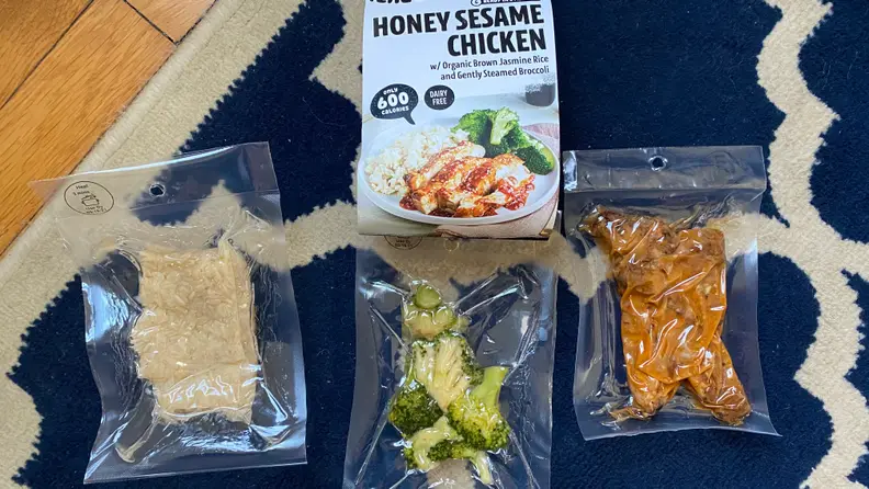 This image displays what's inside a Honey Sesame Chicken premade meal kits, a bag of brown rice, a bag of cooked broccoli, and a bag of sesame chicken.
