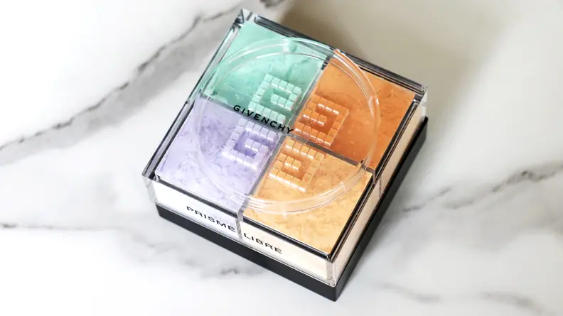 A jar of powder sits on a marble background with its lid off to reveal green, orange, peach, and lavender colors.