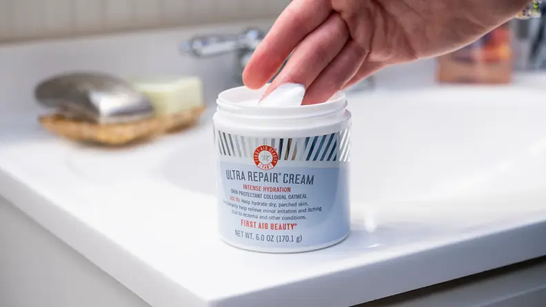 A person's hand dipping into a tub of face moisturizer sitting on a bathroom sink.