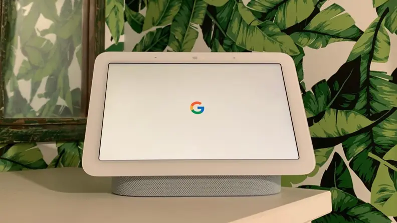 A white Google Nest Hub sitting on a light wood countertop with a tropical plant wallpaper covering the wall in the background.