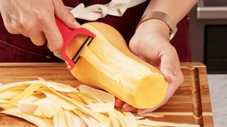 Person using Y-peeler tool to remove skin from butternut squash on top of wooden cutting board next to pile of skin.