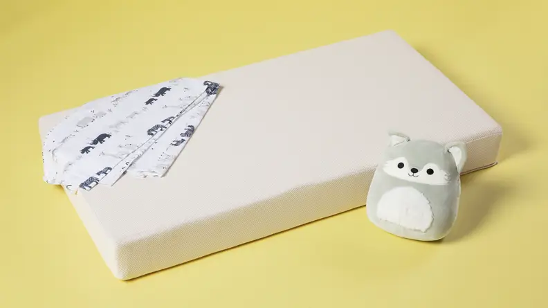 A Colgate Eco Classica III crib mattress on a yellow background with a baby blanket and a stuffed owl.
