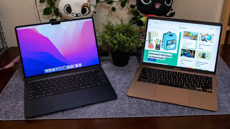 MacBook Air 13 M2 laptop computer in Midnight color with display colorful screensaver on screen next to the MacBook Air 15 M2 laptop computer in rose gold color with browser open on screen Black MacBook Air and rose gold MacBook Air 15 laptops side by side on top of blue cloth in front of plush stuffed animals and potted plant..