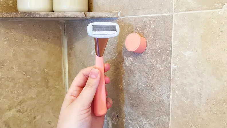 A hand holding a pink Athena Club Razor in front of its wall mount, which is stuck to the shower wall.