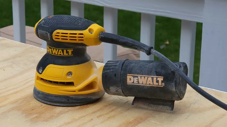 A dust-dust covered Dewalt random orbital sander sits on top of a smooth plank of wood.