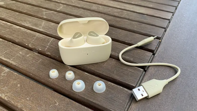 The gold beige Jabra Elite 5 in their open case with included USB-C to USB-A charge cable and extra ear tips on a wooden table.