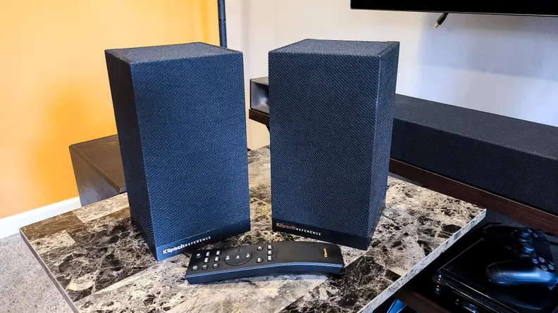 A pair of all-black speakers wrapped in acoustic fabric sit in front of a black, rubberized remote with white-lettered buttons that include subwoofer and surround sound control.