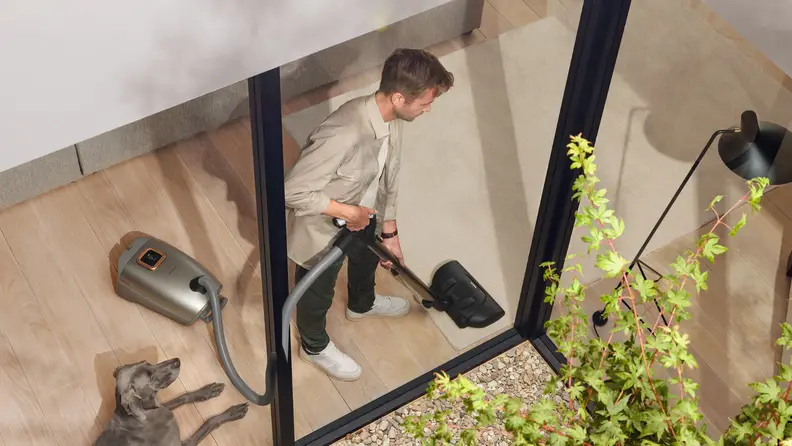 A man vacuums inside with a gray dog watching