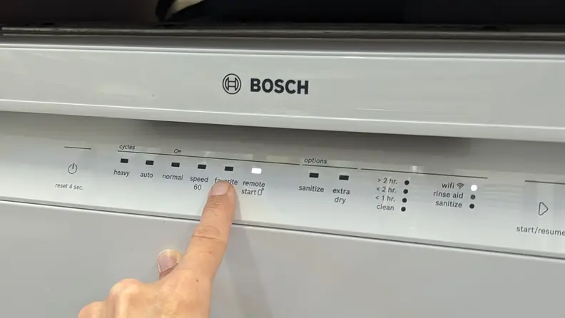 A finger points to the "Favorite" cycle button on the front-facing control panel of the Bosch 100 Series SHE3AEM2N dishwasher.