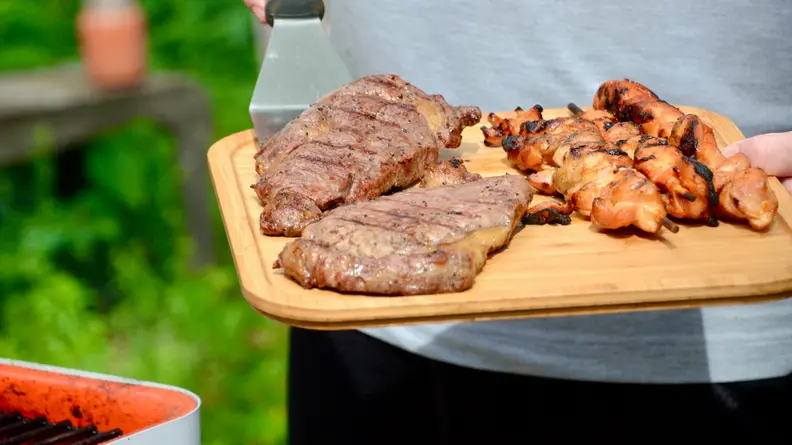A person is holding the bamboo food tray with two pieces of steak and three skewers of chicken on it.