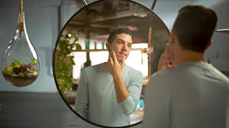 Hyram Yarbro, the founder of Selfless by Hyram, looking into a large round mirror and touching his face with his hand.
