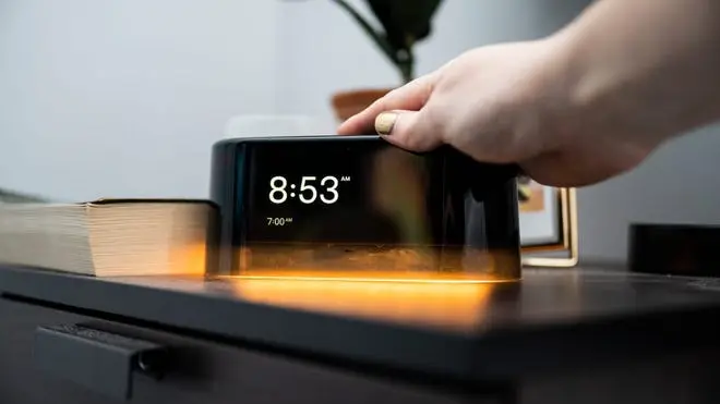 Person touching the top of glowing alarm clock next on bedside table.