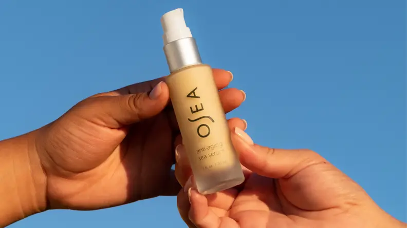 Two hands holding out a small pump bottle of skincare in front of a blue sky.