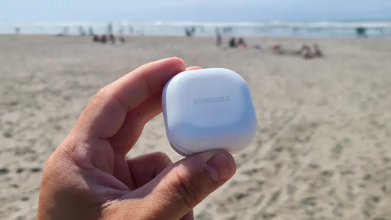 Samsung Galaxy Buds 2 are held in their white case showing against a sunny beach and mountains.