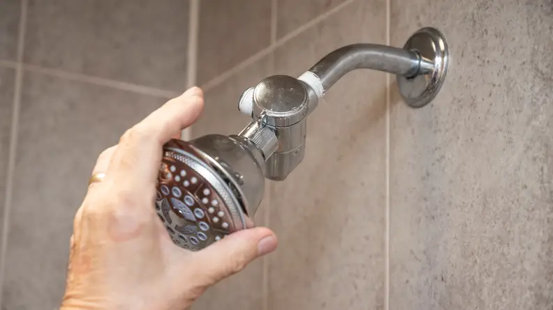 A hand reaches around a silver Hydroluxe showerhead mounted in a shower.