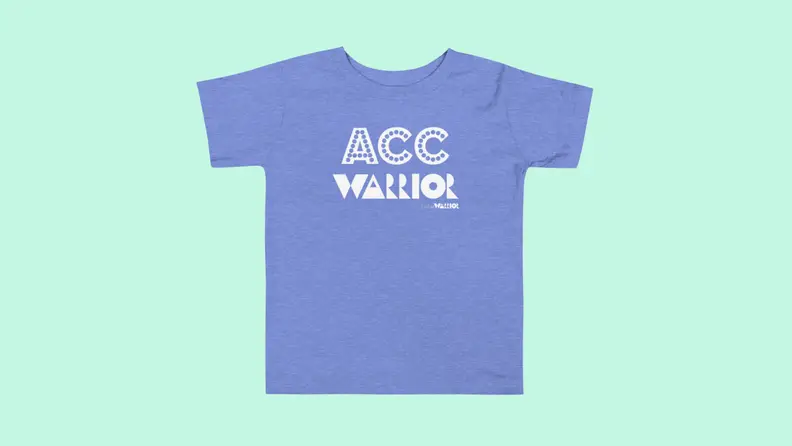 A blue Littlest Warrior T-shirt on a green background.